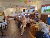 Seniorzy siedzą przy stołach, mają twarze skierowne w stronę występującej osoby